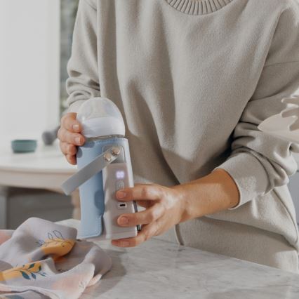 Žena drží v rukách prenosný ohrievač na detská fľaška s fľaškou vo vnútri. Ohrievač je modrý, má uško a displej. Udalosť sa odohráva v kuchyni na kuchynskej linke.