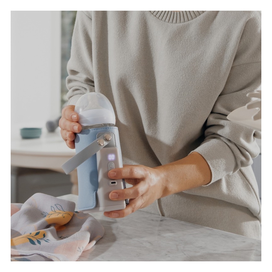 Žena drží v rukách prenosný ohrievač na detská fľaška s fľaškou vo vnútri. Ohrievač je modrý, má uško a displej. Udalosť sa odohráva v kuchyni na kuchynskej linke.