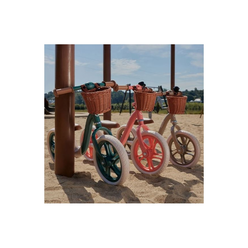 Tri odrážadlá s prútenými košíkmi stoja na piesku pri detskom ihrisku. Dve sú zelené a ružové, jedna hnedá. Odražadlá sú opreté o drevený stĺpik.