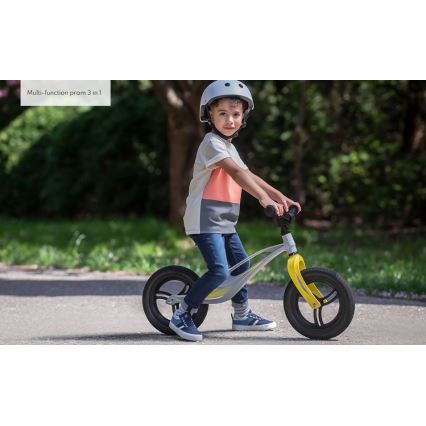Dieťa jazdí na odrážadle šedo-žltej farby po asfaltovej ceste v parku. Dieťa má na hlave prilbu. Text na obrázku: Multi-function pram 3 in 1.