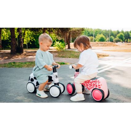 Dve malé deti sedia na odrážadlách. Dieťa na bielom odrážadle s čiernobielym vzorom má na sebe svetlomodré šaty. Druhé dieťa sedí na ružovom odrážadle s leopardím vzorom a má na sebe biely sveter a béžové nohavice. Obe deti sa nachádzajú na asfaltovanej ploche v parku.