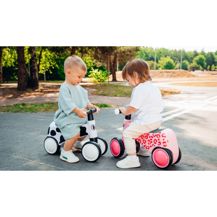 Dve malé deti sedia na odrážadlách. Dieťa na bielom odrážadle s čiernobielym vzorom má na sebe svetlomodré šaty. Druhé dieťa sedí na ružovom odrážadle s leopardím vzorom a má na sebe biely sveter a béžové nohavice. Obe deti sa nachádzajú na asfaltovanej ploche v parku.