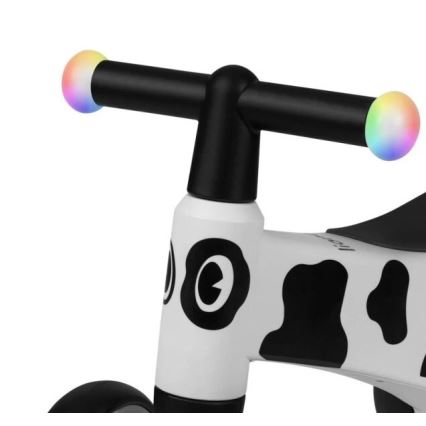 Detské odrážadlo s bielym rámom a čierno-bielym vzorom kravy je zobrazené zblízka. Ovládací riadidlá majú na koncoch farebne blikajúce svetielka. Rám je prevažne biely s čiernymi škvrnami.