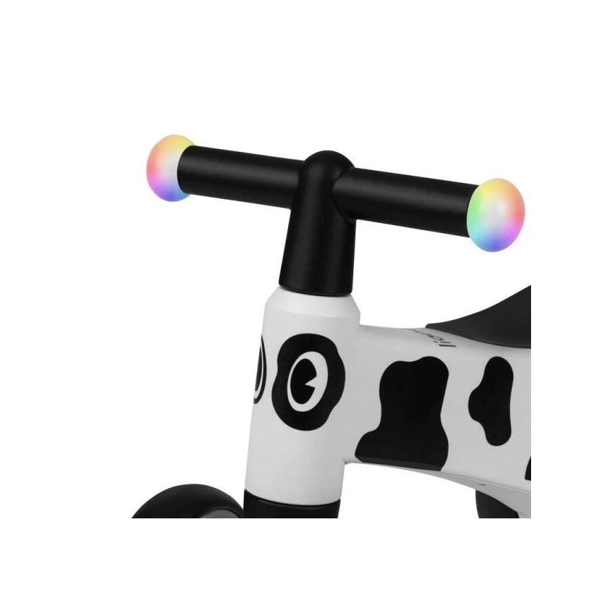 Detské odrážadlo s bielym rámom a čierno-bielym vzorom kravy je zobrazené zblízka. Ovládací riadidlá majú na koncoch farebne blikajúce svetielka. Rám je prevažne biely s čiernymi škvrnami.