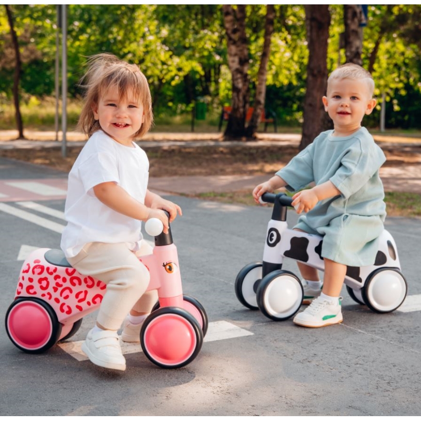 Dve malé deti sa vo vonkajšom prostredí preháňajú na odrážadlách. Dieťa na ružovom odrážadle s leopardím vzorom má na sebe biely top a béžové nohavice. Dieťa na čierno-bielej odrážadle s vzorom kravy má na sebe svetlomodré tričko. Odražadlá majú tri kolesá.