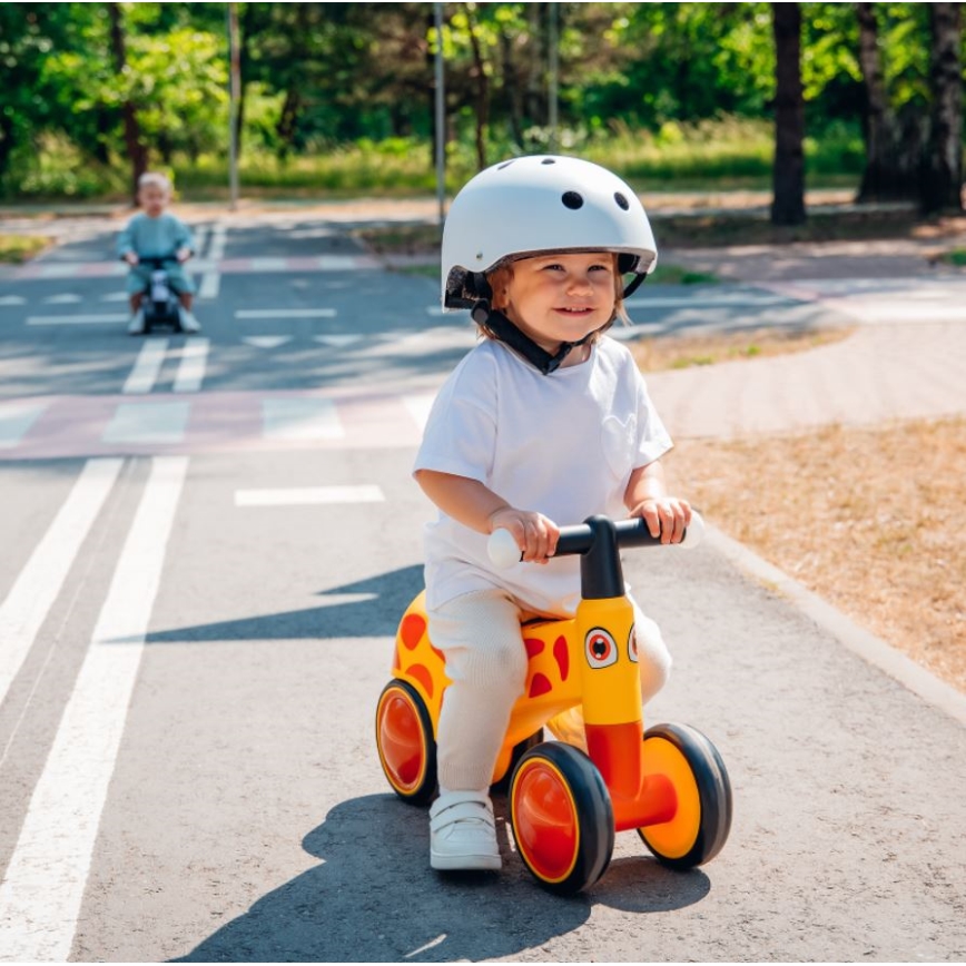 Dieťa v prilbe šoféruje žlté odrážadlo v tvare žirafy po chodníku v parku. V pozadí je ďalšie dieťa na podobnom odrážadle.