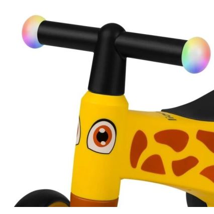 Detské odrážadlo tvaru žirafy s čiernou riadidlami a farebnými svetlami na koncoch. Odrážadlo je žlté s hnedými škvrnami.