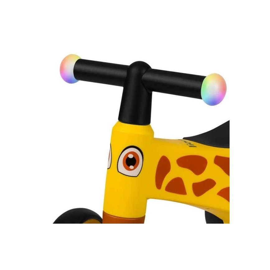 Detské odrážadlo tvaru žirafy s čiernou riadidlami a farebnými svetlami na koncoch. Odrážadlo je žlté s hnedými škvrnami.