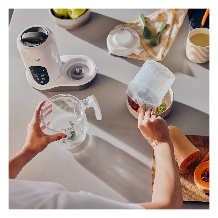 Osoba prekladá uvarenú zeleninu z nádoby do misky. Používa pri tom kuchynský robot Lionelo. Na stole sú ďalšie ingrediencie a kuchynské pomôcky.