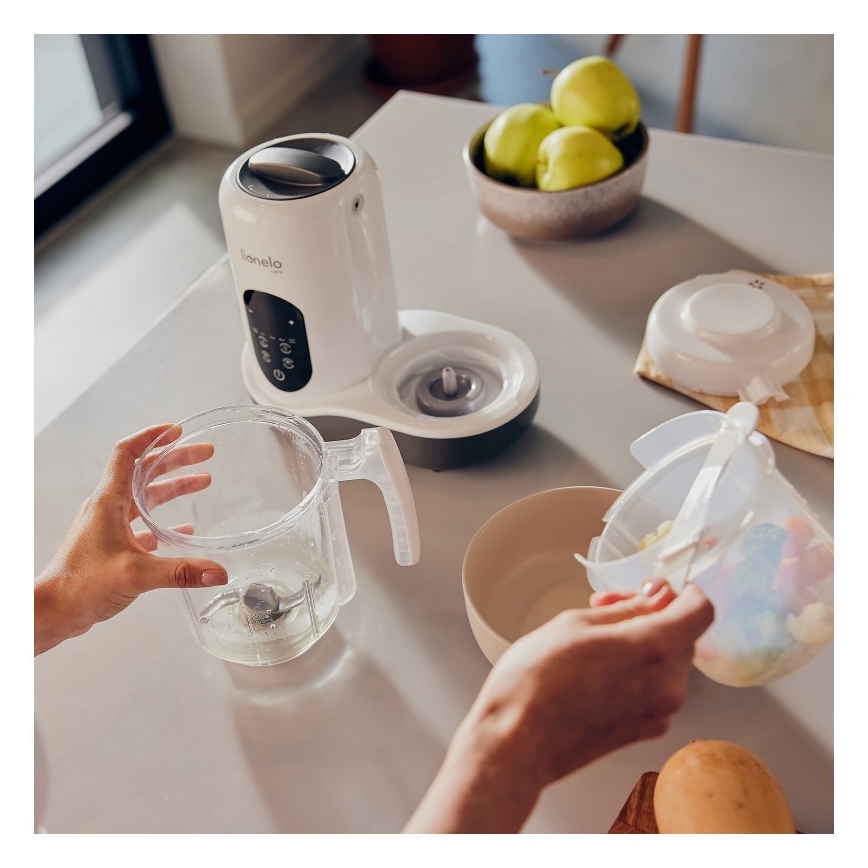 Osoba pripravuje jedlo v kuchynskom robote značky Lionelo. Do nádoby robota vkladá suroviny z plastovej nádobky. Na stole sa nachádza miska s jablkami a ďalšie kuchynské náčinie.