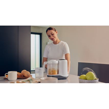 Žena umiestňuje na kuchynský stôl mixér na prípravu detskej výživy obsahujúci pomixovanú oranžovú zeleninu. Na stole sú ďalšie misky, hrnček, zelenina a ovocie.