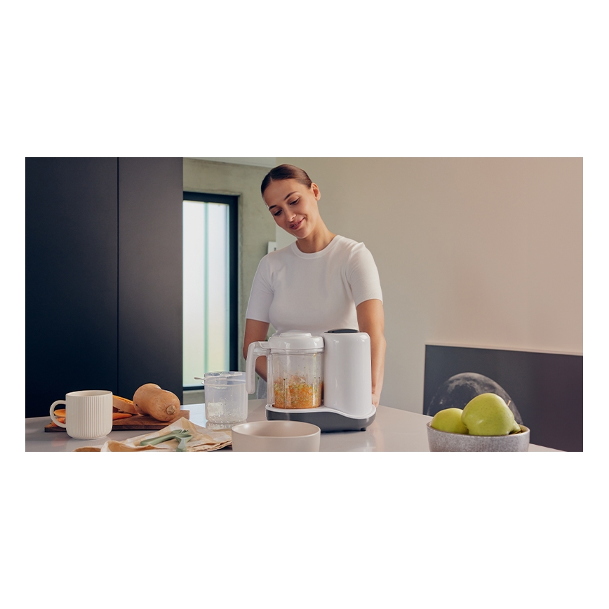 Žena umiestňuje na kuchynský stôl mixér na prípravu detskej výživy obsahujúci pomixovanú oranžovú zeleninu. Na stole sú ďalšie misky, hrnček, zelenina a ovocie.