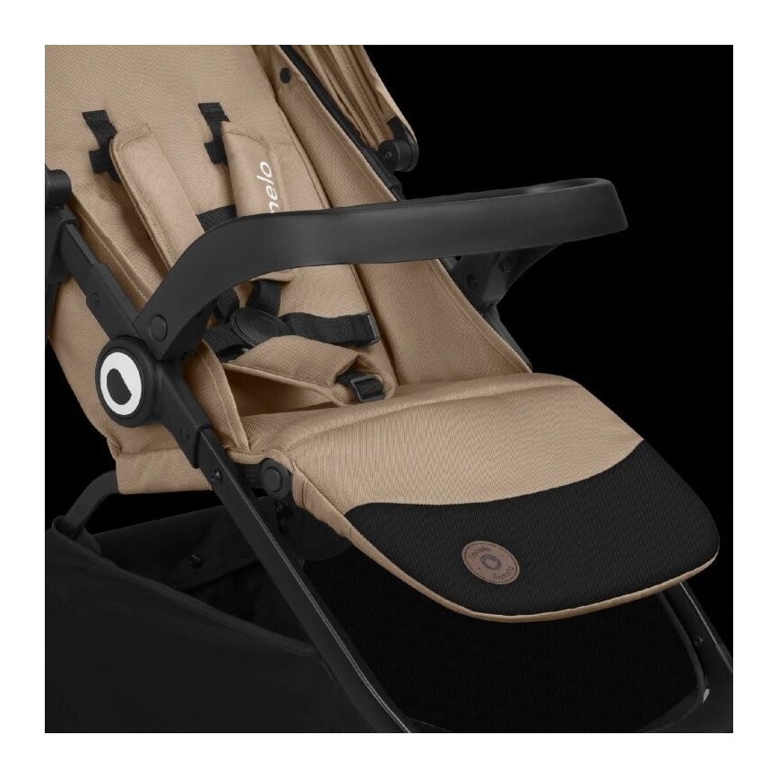 Kočík je sklopený do polohy na ležanie. Má béžové čalúnenie a čierne plastové prvky, vrátane opierky rúk. Je zobrazený na čiernom pozadí.