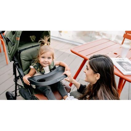 Dieťa sedí v zelenom športovom kočíku s čiernou podnosovou táckou s nápisom „Lionelo“. Matka sa usmieva a pridržiava podnos. Kontext: vonkajšie posedenie s oranžovým stolíkom.