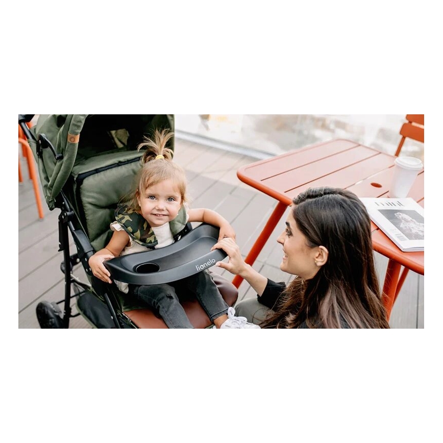 Dieťa sedí v zelenom športovom kočíku s čiernou podnosovou táckou s nápisom „Lionelo“. Matka sa usmieva a pridržiava podnos. Kontext: vonkajšie posedenie s oranžovým stolíkom.