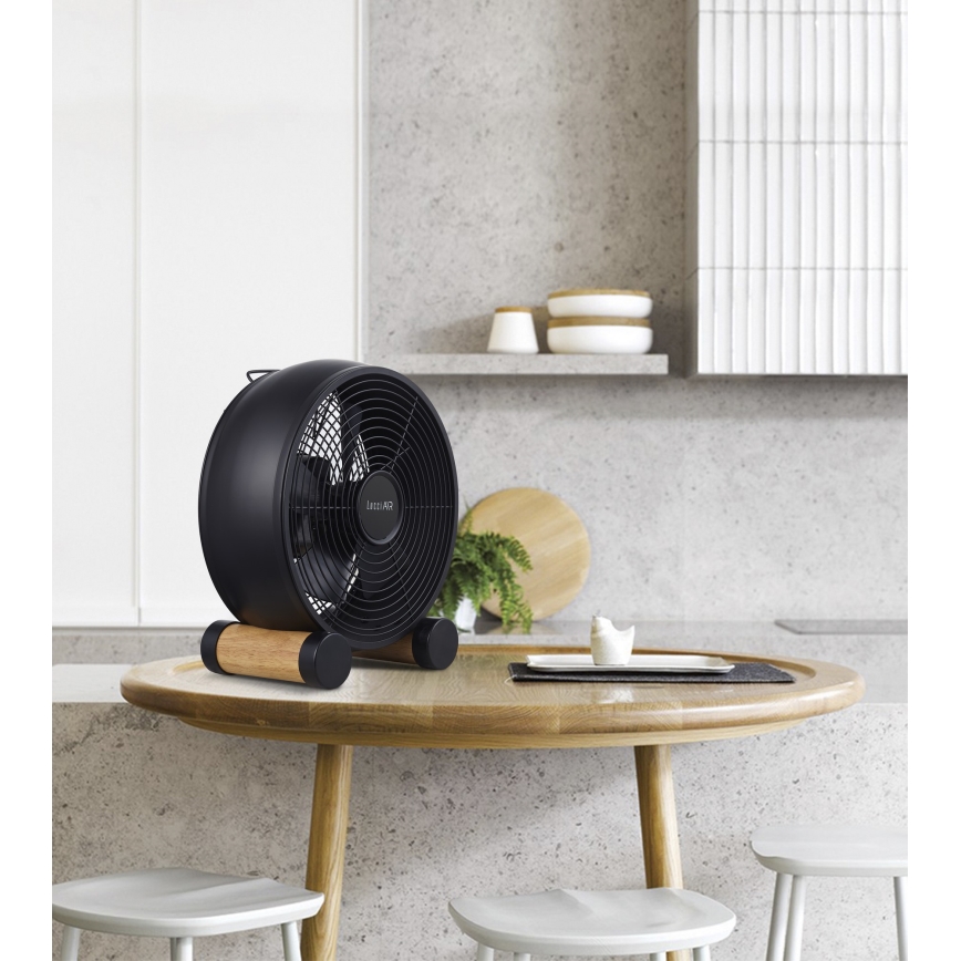 Čierny stolový ventilátor s drevenými nožičkami stojí na drevenom stole v kuchyni. Ventilátor je zapnutý. Na stole je aj malá keramická figúrka a podnos.