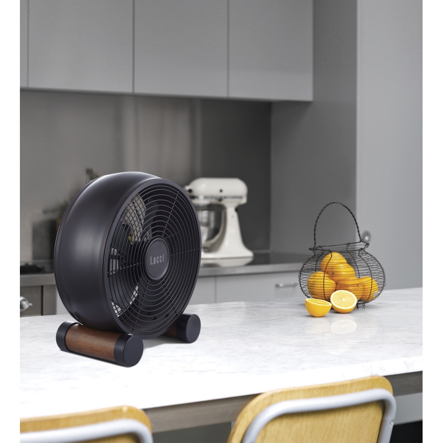 Čierny stolový ventilátor Locii stojí na drevenom podstavci na kuchynskej linke. V pozadí sa nachádza kuchynská linka s kuchynskými spotrebičmi a košíkom s pomarančmi a citrónmi.