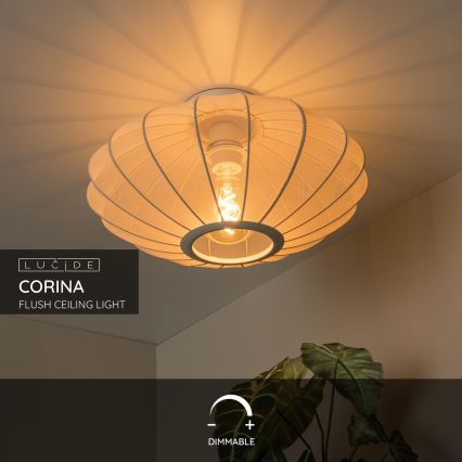 Svietidlo Corina svieti. Ide o nástenné svietidlo s bielym tienidlom z látky. Nachádza sa v interiéri, pri stene s rastlinou. Text: LUCIDE CORINA FLUSH CEILING LIGHT DIMMABLE.