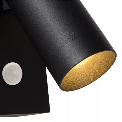 Čierna nástenná lampa s valcovým tienidlom so zlatým vnútorným povrchom svieti. Lampa je pripevnená na stene a obsahuje zabudovaný snímač.