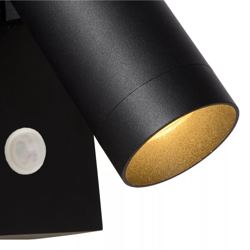 Čierna nástenná lampa s valcovým tienidlom so zlatým vnútorným povrchom svieti. Lampa je pripevnená na stene a obsahuje zabudovaný snímač.