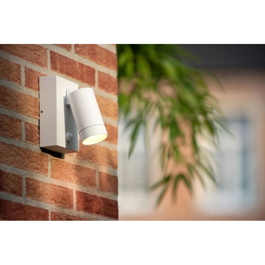 Biela vonkajšia lampa s detektorom pohybu je pripevnená na tehlovej stene. Svieti. Na pozadí je zeleň.
