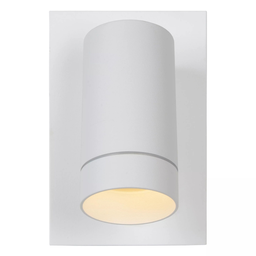 Biela nástenná lampa s valcovým tienidlom svieti. Lampa je pripevnená na obdĺžnikovú bielu základňu.