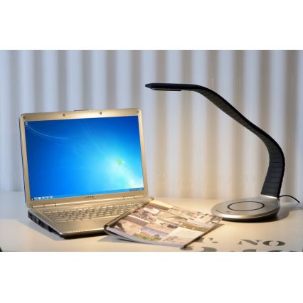 Stolná lampa s ohnutým ramenom svieti na notebook a časopis položené na stole.