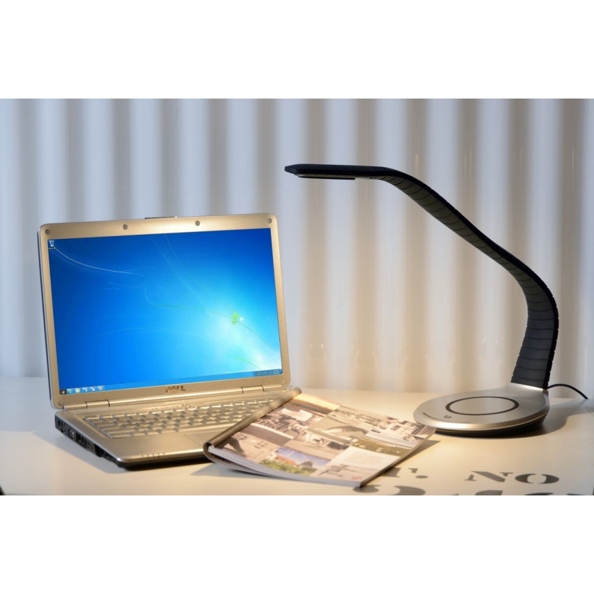 Stolná lampa s ohnutým ramenom svieti na notebook a časopis položené na stole.