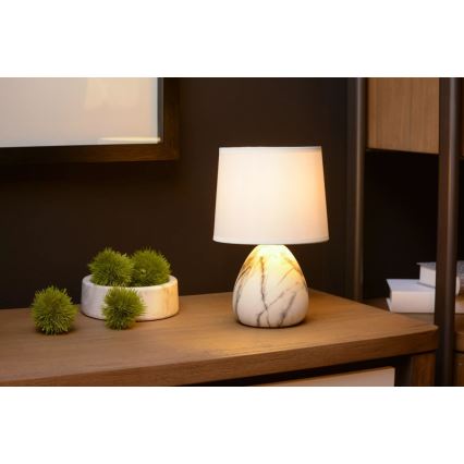 Stolová lampa s bielym tienidlom a mramorovým podstavcom svieti na drevenom stole. Na stole je aj miska s umelými zelenými guličkami. V pozadí je tmavá stena a časť nábytku.
