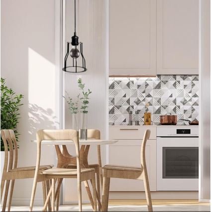 Závesná lampa visí nad jedálenským stolom v kuchyni. Lampa je čierna, kovová s klietkou okolo žiarovky. Stôl je obklopený drevenými stoličkami. Kuchyňa má svetlé drevené skrinky a geometricky vzorované obklady.