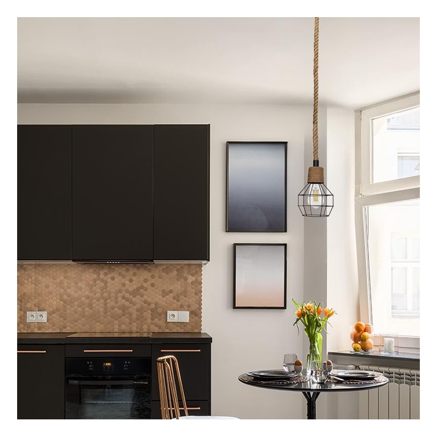 Závesné svietidlo visí v kuchyni. Je zavesené na šnúre a má kovový kôš. Svietidlo sa nachádza nad okrúhlym stolom s jedálenským servisom. V pozadí sú tmavé kuchynské skrinky a svetlá stena s obrazmi.