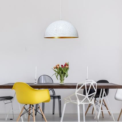 Biela visiaca lampa s perforáciami a zlatým vnútorným okrajom visí nad jedálenským stolom. Stôl je obklopený rôznymi stoličkami. Na stole je váza s tulipánmi.