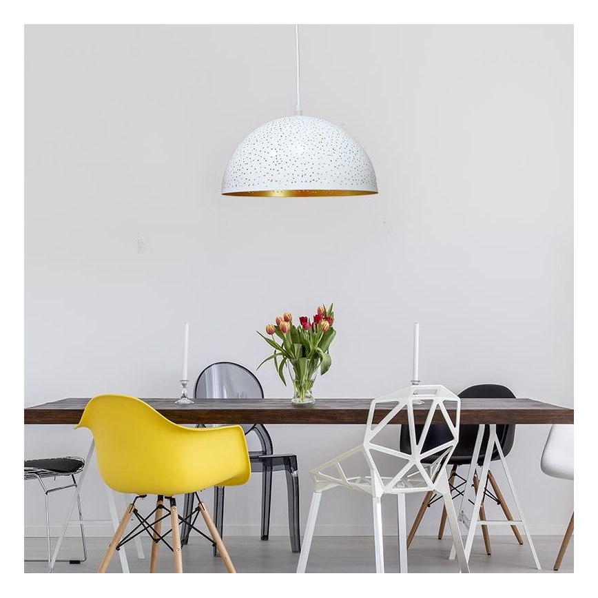 Biela visiaca lampa s perforáciami a zlatým vnútorným okrajom visí nad jedálenským stolom. Stôl je obklopený rôznymi stoličkami. Na stole je váza s tulipánmi.