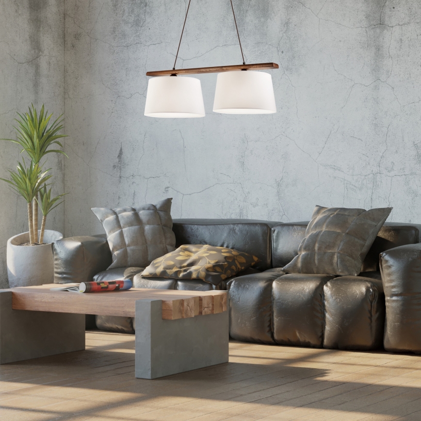 Dvojité závesné svietidlo visí nad koženou sedačkou v industriálne ladenom interiéri. Svietidlo má drevenú konštrukciu a dva biele tienidlá. Na konferenčnom stolíku leží časopis.