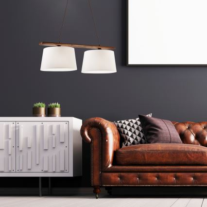 Dvojité závesné svietidlo s bielymi tienidlami a drevenou konštrukciou visí nad hnedou koženou pohovkou. Pohovka je umiestnená pred bielou komodou s geometrickým vzorom. V pozadí je tmavá stena a prázdny rám obrazu.