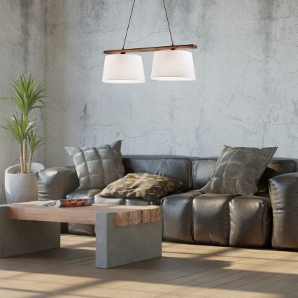 Dvojité závesné svietidlo visí nad koženou sedačkou v industriálne ladenom interiéri. Svietidlo má drevenú konštrukciu a dva biele tienidlá. Na konferenčnom stolíku leží časopis.