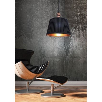 Čierna závesná lampa s medeným detailom visí nad čiernym koženým kreslom a podnožkou s drevenými prvkami. Kreslo a podnožka stoja na drevenej podlahe pred betónovou stenou.