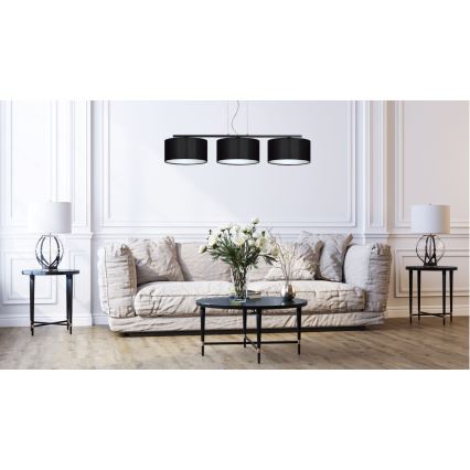 Trojramenné závesné svietidlo s čiernymi tienidlami visí nad pohovkou v obývacej izbe. Svietidlo je umiestnené nad nízkym kávovým stolíkom, na ktorom je váza s kvetmi. Po stranách pohovky sú umiestnené dve malé stolíky s lampami.