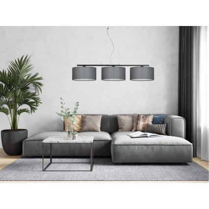 Trojramenné závesné svietidlo s tmavosivými tienidlami visí nad sivou rohovou sedačkou v obývacej izbe. Na konferenčnom stolíku pred sedačkou sú dekorácie.