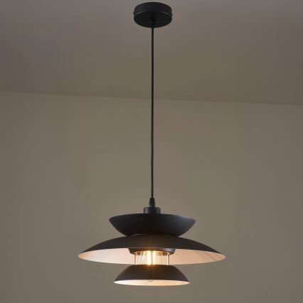 Čierna závesná lampa s dvoma tienidlami visí zo stropu v svetlohnedej miestnosti. Svieti vnútorná žiarovka.