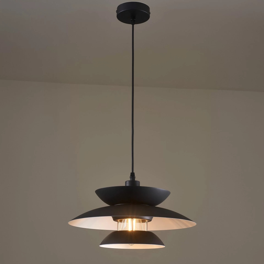 Čierna závesná lampa s dvoma tienidlami visí zo stropu v svetlohnedej miestnosti. Svieti vnútorná žiarovka.
