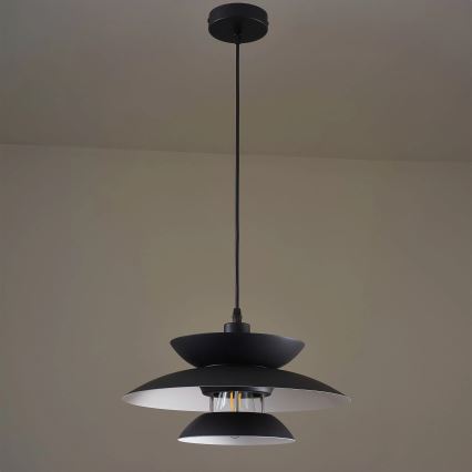 Čierna závesná lampa s tromi tienidlami visí z stropu v svetlohnedej miestnosti. Spodné tienidlo je najväčšie. V strede je viditeľná žiarovka.