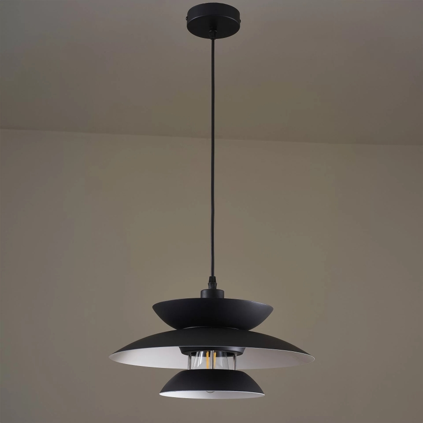Čierna závesná lampa s tromi tienidlami visí z stropu v svetlohnedej miestnosti. Spodné tienidlo je najväčšie. V strede je viditeľná žiarovka.