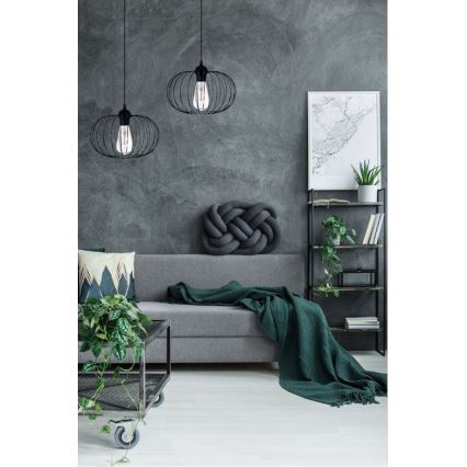 Dve čierne kovové závesné lampy svietia v obývacej izbe. Sivá sedačka s vankúšmi a zelenou dekou je doplnená o stolík a regál s rastlinami.