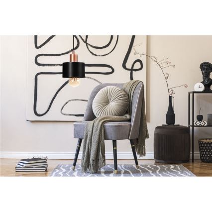 Závesné svietidlo s čiernym tienidlom a medeným detailom visí nad sivým kreslom s vankúšom a prehozom. Kreslo stojí v miestnosti s abstraktným obrazom na stene a doplnkami ako stolík a váza.