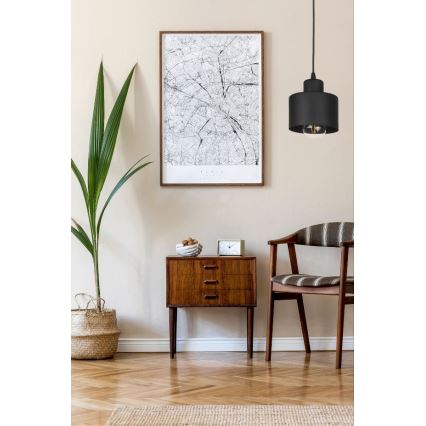 Svietidlo visí v interiéri. Je umiestnené nad dreveným nočným stolíkom s tromi zásuvkami, vedľa kresla a rastliny v koši. Na stene za ním visí obraz.