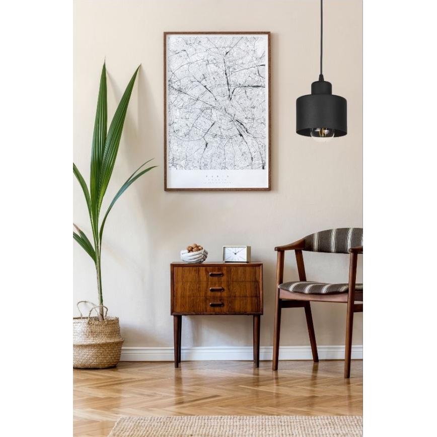 Svietidlo visí v interiéri. Je umiestnené nad dreveným nočným stolíkom s tromi zásuvkami, vedľa kresla a rastliny v koši. Na stene za ním visí obraz.