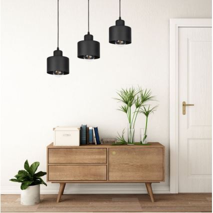 Tri čierne závesné lampy svietia nad drevenou komodou. Komoda stojí v miestnosti s bielymi stenami a dverami. Na komode sú knihy a rastliny.