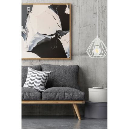 Biela kovová lampa visí v interiéri. Svietidlo je umiestnené nad sivou pohovkou s vankúšmi. Pozadie tvorí betónová stena a obraz.