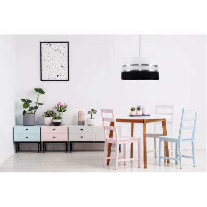 Závesné svietidlo svieti nad jedálenským stolom s tromi stoličkami. Stolík a stoličky sú svetlé, drevené. Vedľa stola je komoda s tromi zásuvkami v pastelových farbách.